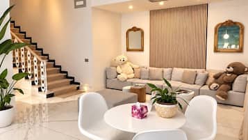 A modern living room with a staircase, a sofa set, two white chairs, a round table, and decorative elements like a mirror and plants.
