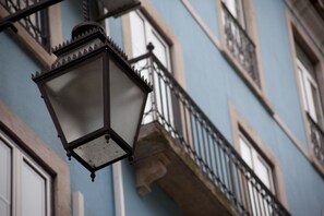 A close-up of a hexagonal street lamp with a frosted glass shade, attached to a building with a blue facade and black wrought iron balcony railings.