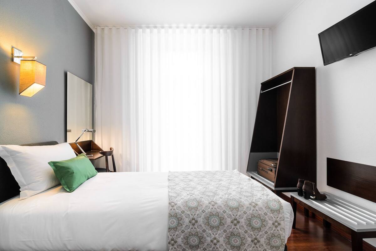 Une chambre d’hôtel avec un lit, une table de chevet, un téléviseur fixé au mur et une grande fenêtre avec des rideaux.
