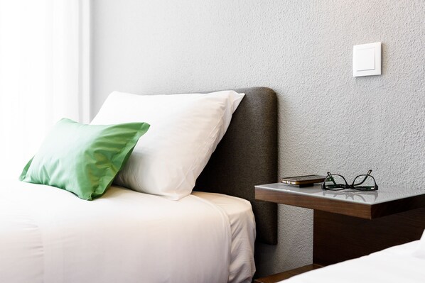 A neatly made bed with white and green pillows, a bedside table with a book and glasses, and a textured wall.