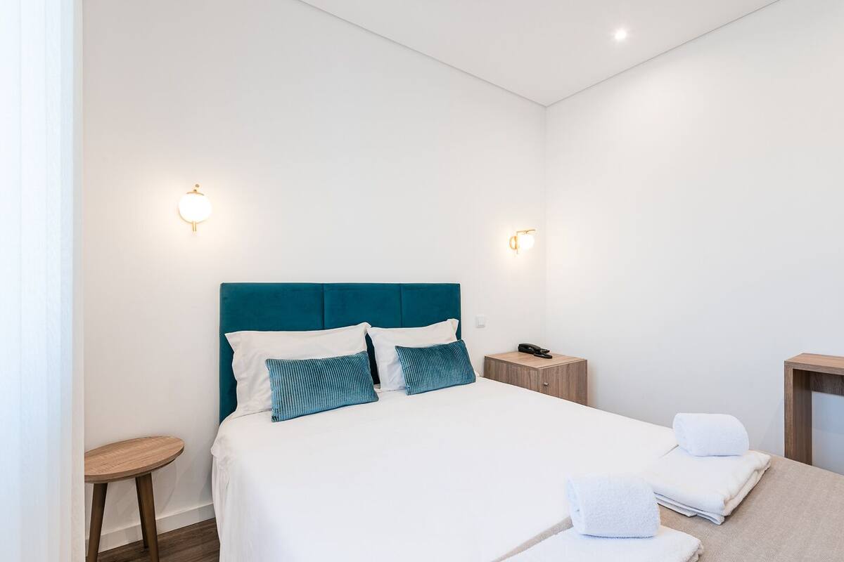 A modern hotel room with a teal headboard, white bedding, and two wall-mounted lights.