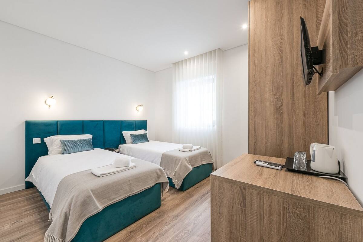 A hotel room with two beds, a wooden desk, a flat-screen TV, and a window with curtains.