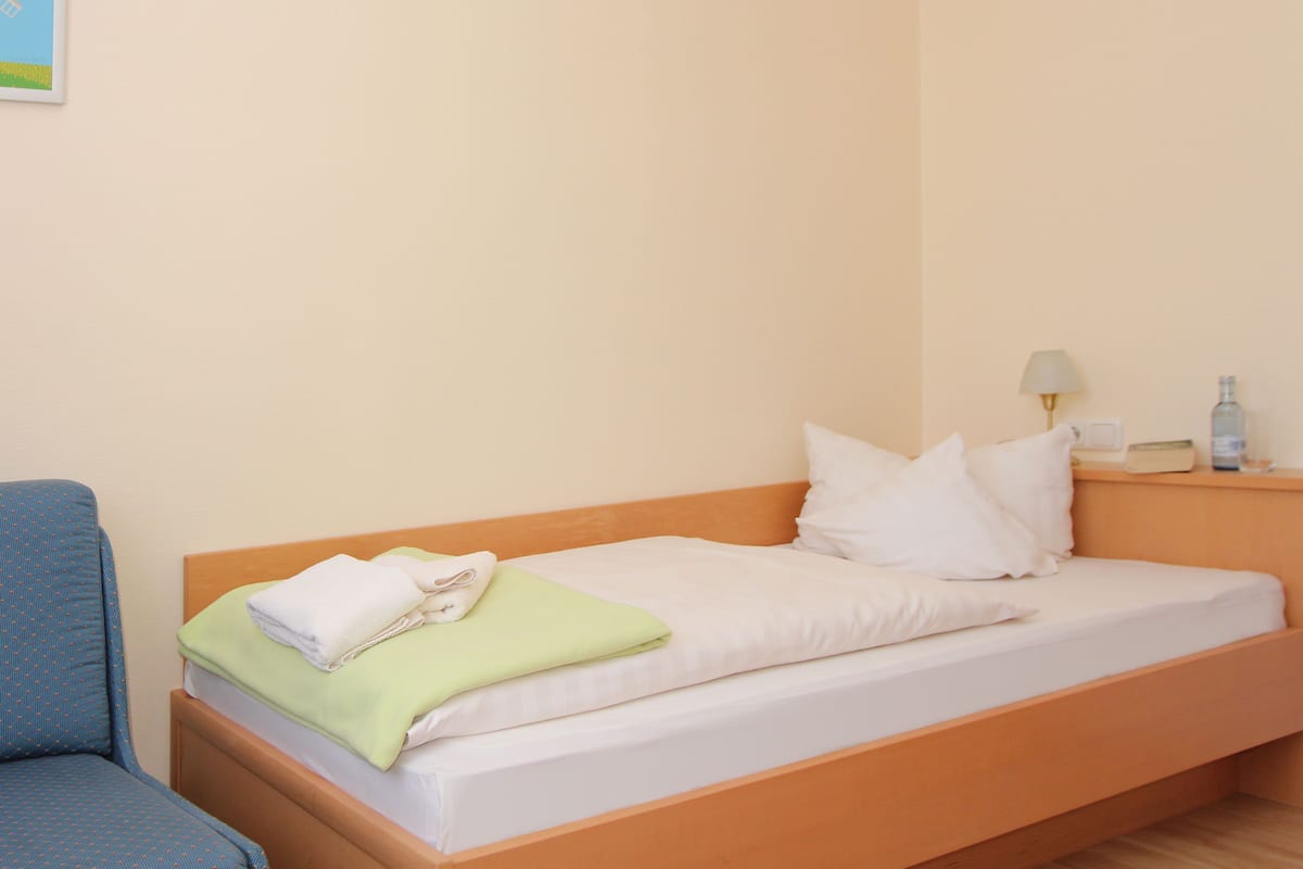 Uma cama de solteiro com roupa de cama branca, um cobertor verde e um travesseiro branco. Há uma mesa de cabeceira com uma luminária e uma garrafa. Uma cadeira azul está visível à esquerda da cama.