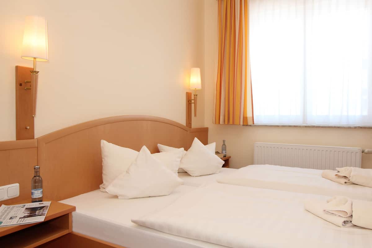 Quarto de hotel com duas camas, cabeceira de madeira, mesa de cabeceira com uma garrafa e janela com cortinas.
