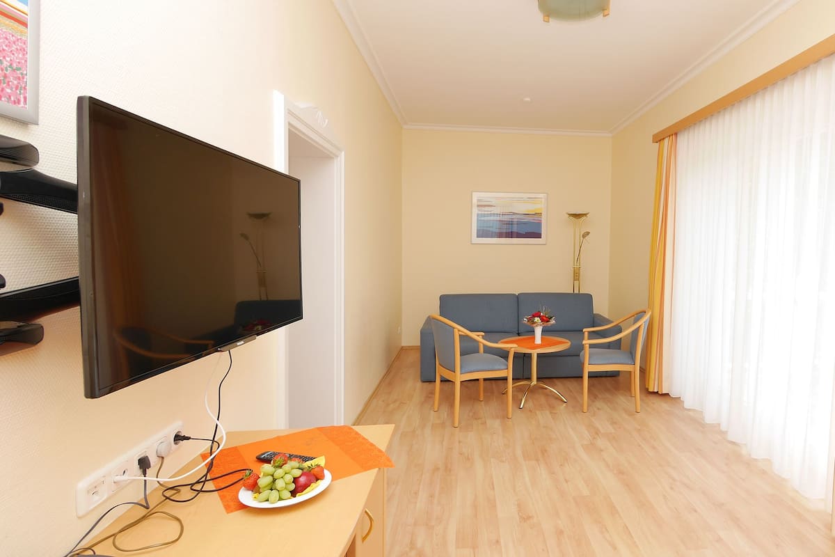 Uma sala de estar com televisão de tela plana, sofá, mesa pequena com frutas e janela com cortinas.