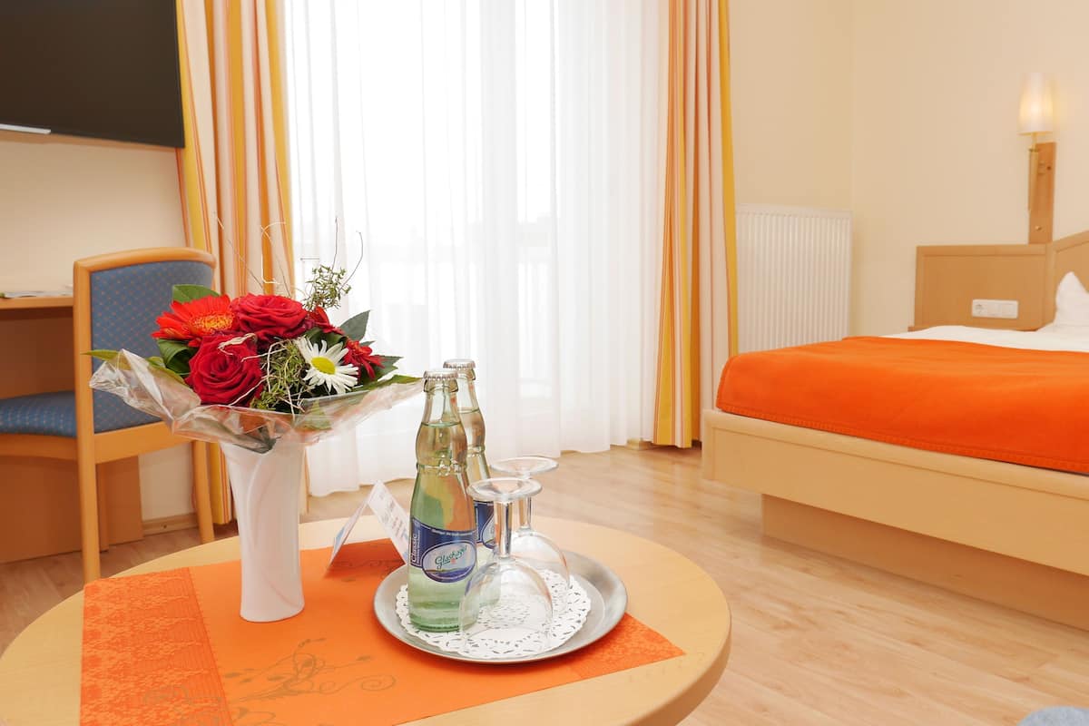 Quarto de hotel com cama, escrivaninha, cadeira, mesa com bandeja, um vaso de flores e uma garrafa de água.
