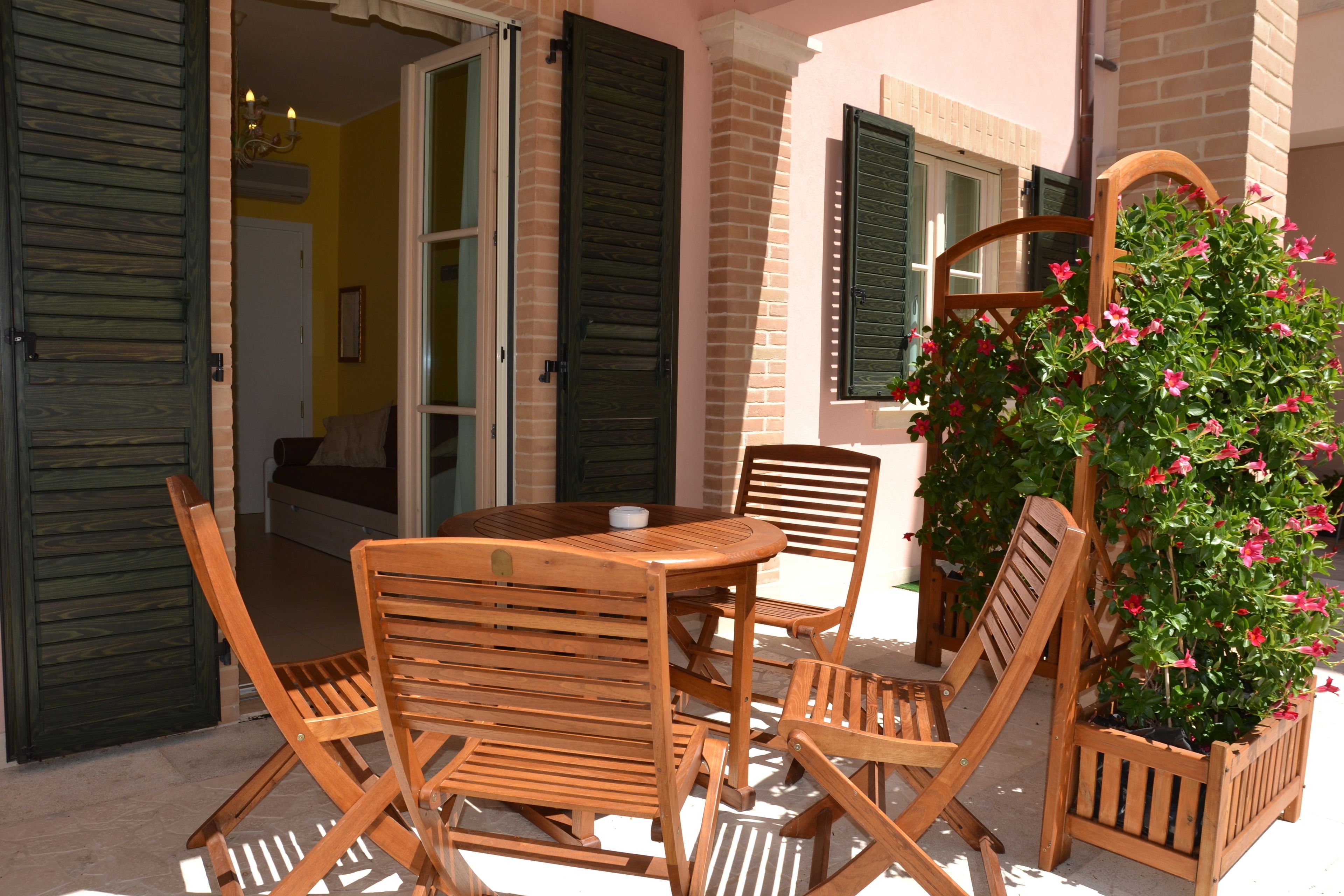 Conjunto de mesa e cadeiras de madeira ao ar livre, em frente a um prédio com persianas abertas e um vaso com flores cor-de-rosa.