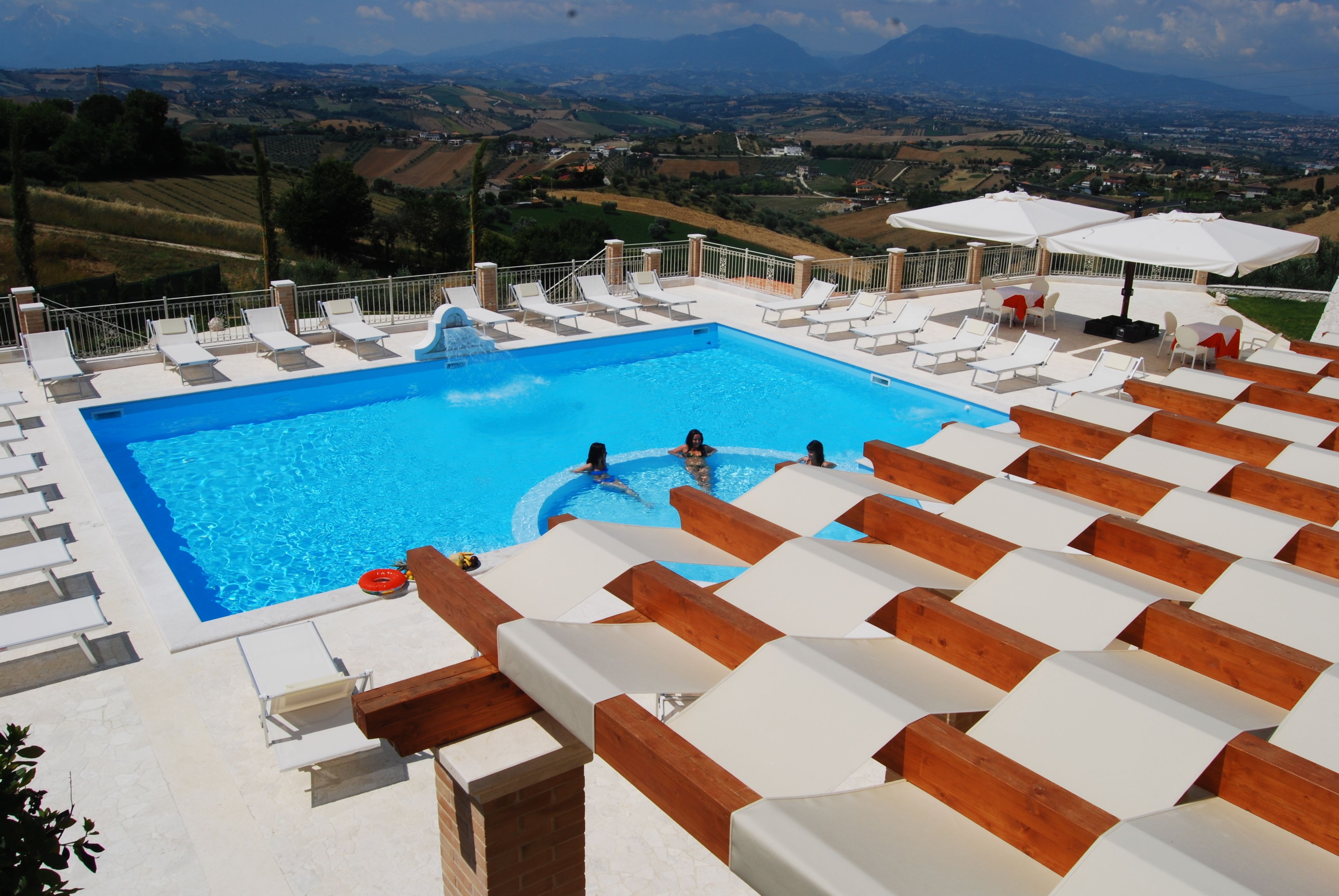 A pool with lounge chairs and umbrellas, surrounded by a wooden deck and a view of the countryside.