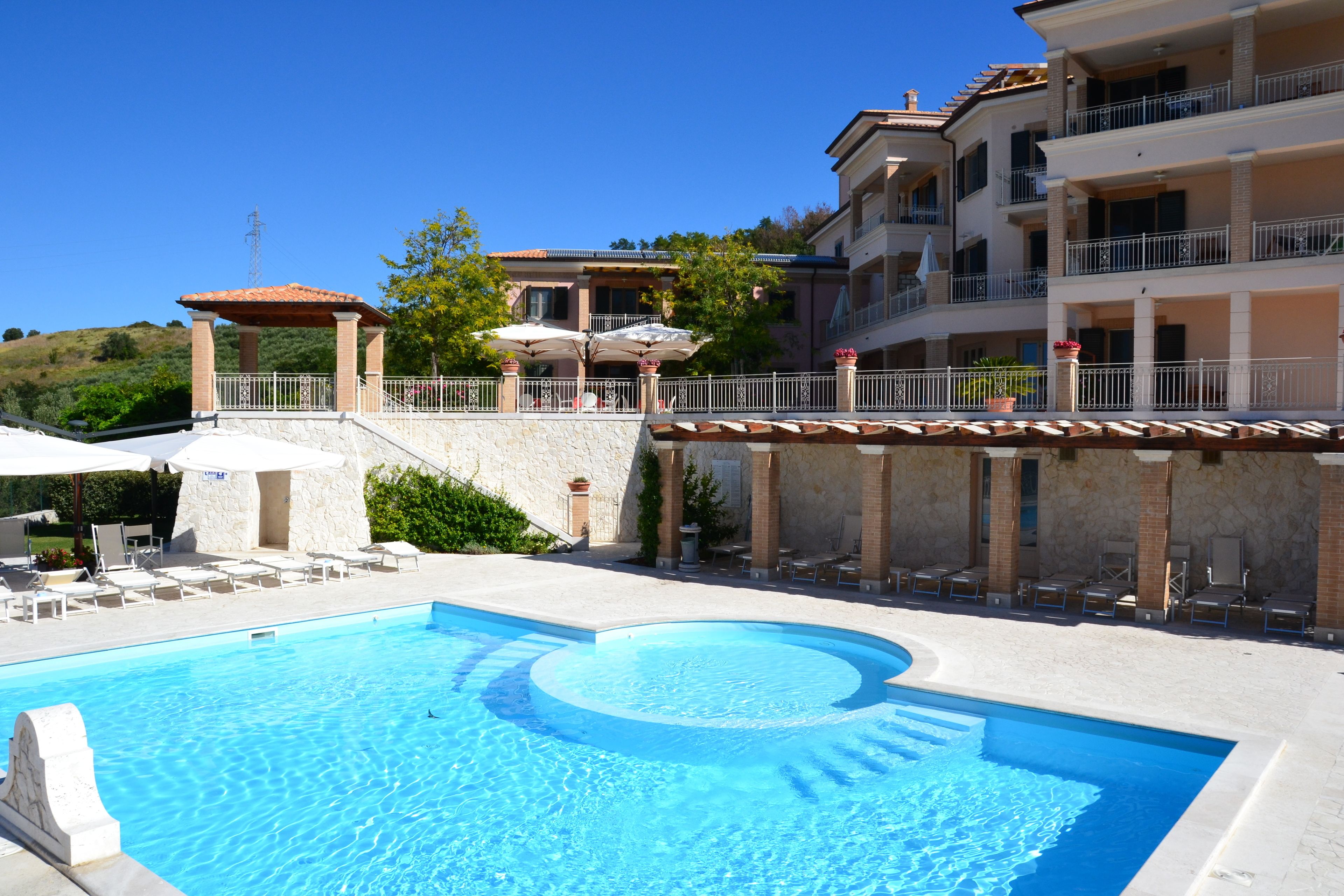 Une piscine entourée de chaises longues et de parasols, le tout bordé d’un bâtiment doté de balcons et d’un mur en pierre.