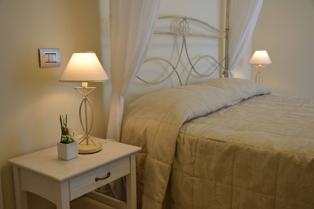 A neatly made bed with a beige quilt, a bedside table with a lamp, and a small potted plant.