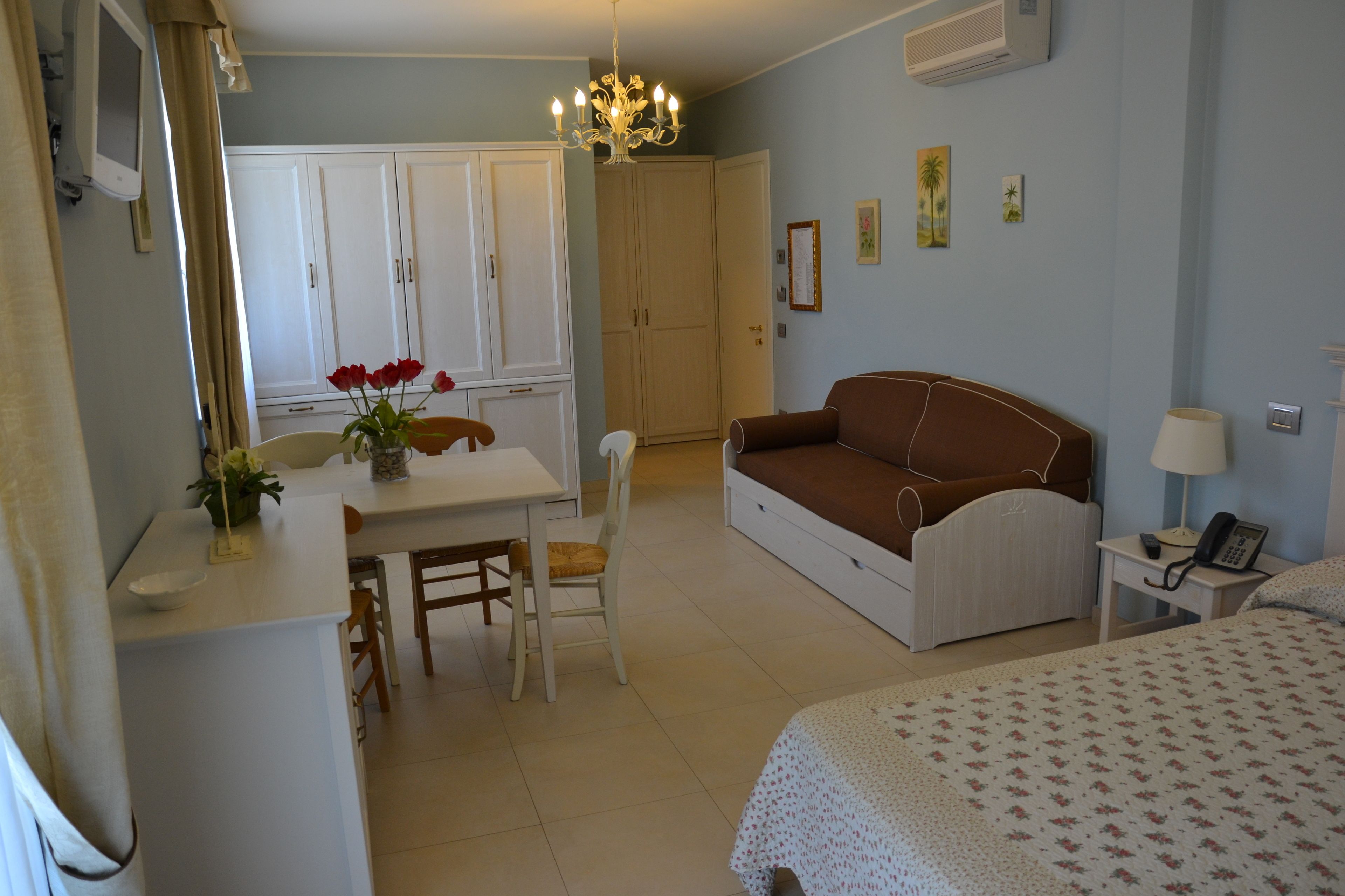 A room with a bed, a sofa, a dining table with chairs, a chandelier, and a wardrobe.
