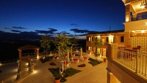 Un patio exterior bien iluminado con varias mesas y sillas, rodeado de plantas en macetas y un pequeño pabellón.