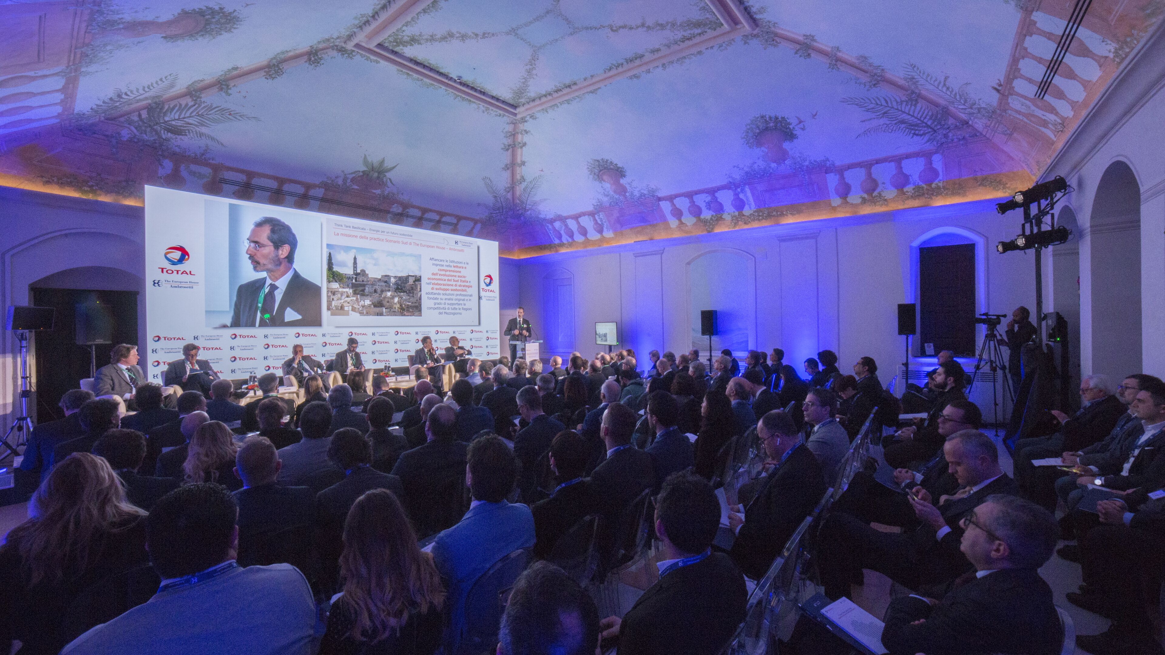 A conference room with a large screen displaying a presentation, a stage with a speaker, and an audience seated at tables.