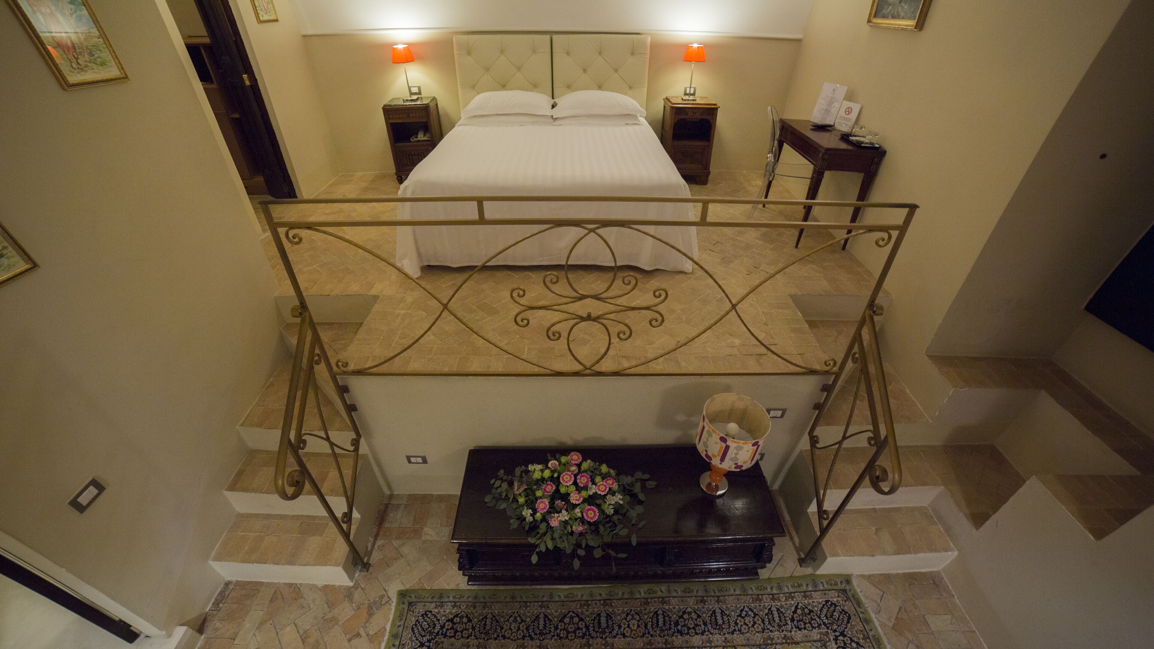 A hotel room with a double bed, two bedside tables, a nightstand, a small table with a vase of flowers, and a decorative wall piece.
