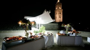 An outdoor event setup with tables covered in white cloths, decorated with flowers and fruit. A white tent is set up in the background, and a tower is visible in the distance.