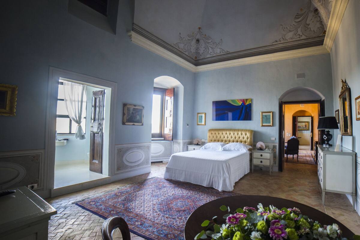 Un dormitorio amplio con una cama grande, una zona de estar con una mesa y sillas, y vista al pasillo.