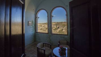 A room with two arched windows offering a view of a rocky landscape. There are two wooden chairs and a small table with a vase.