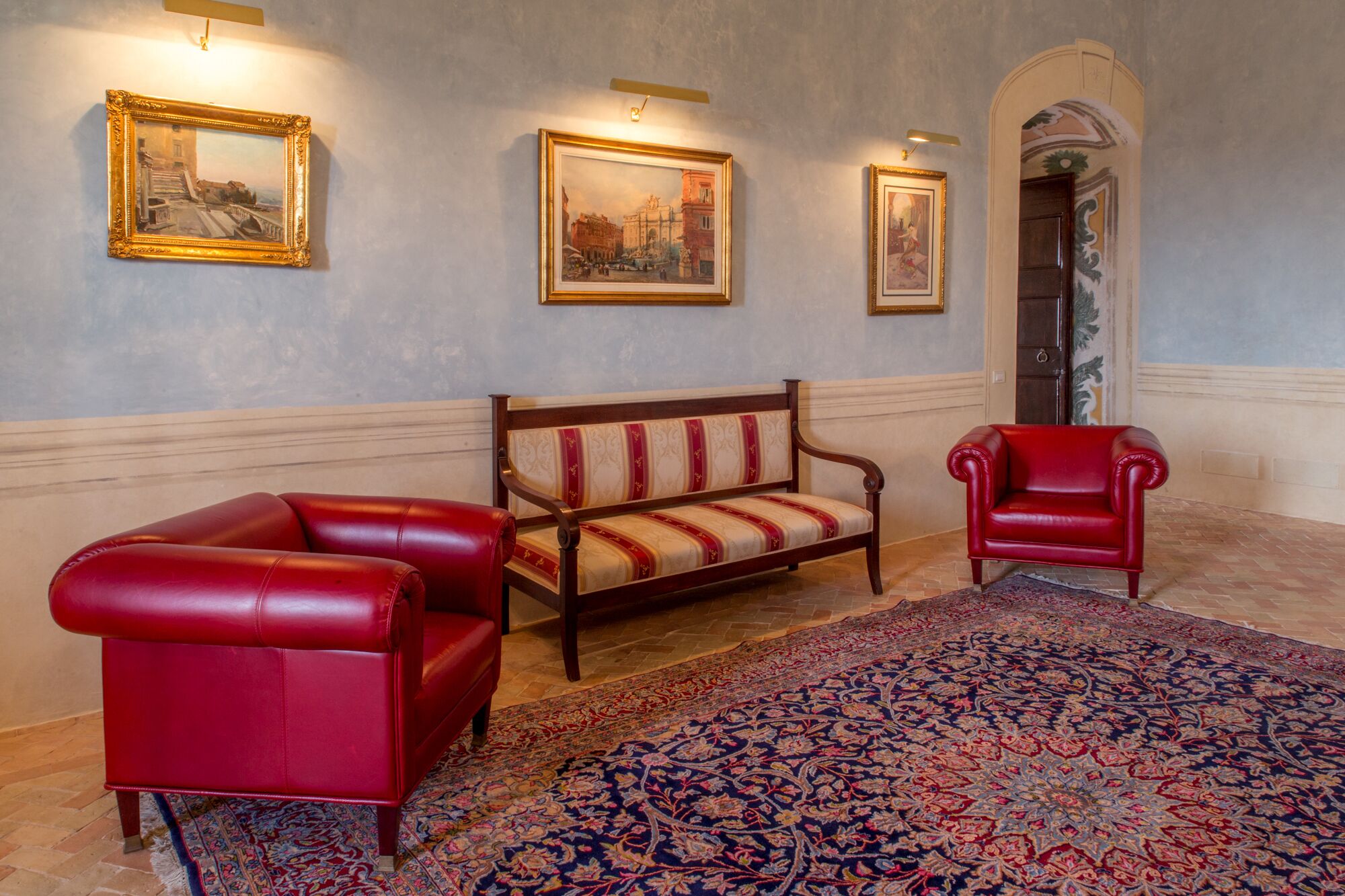 A room with a red leather sofa, a wooden bench with red and beige upholstery, a red armchair, and a patterned rug on the floor.