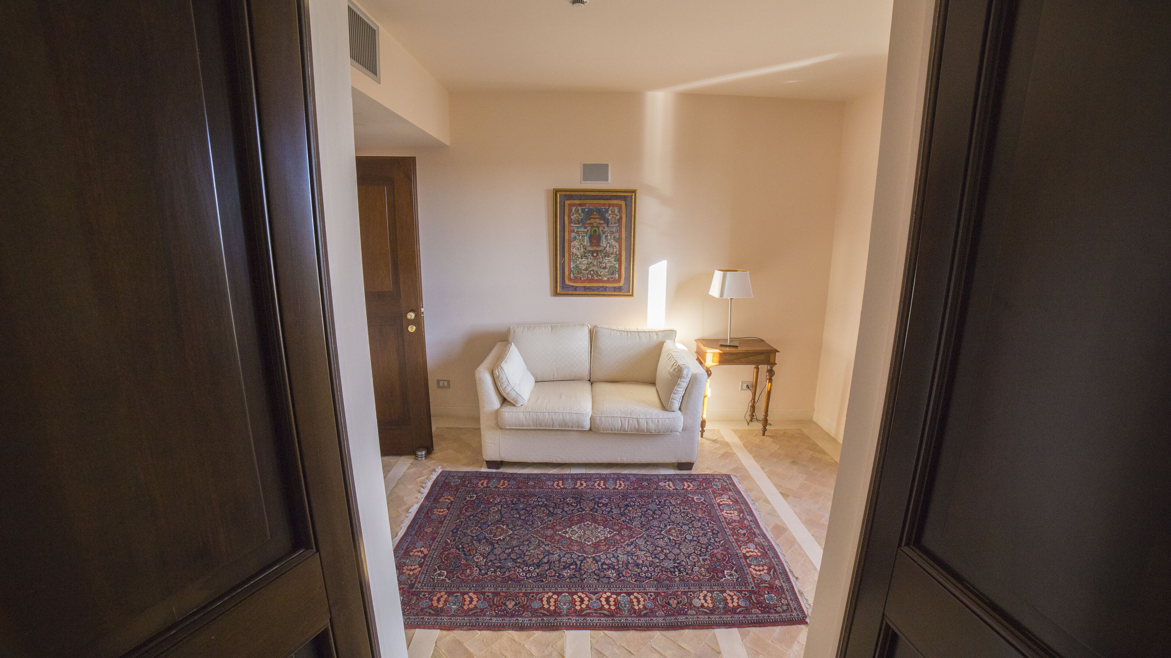 A living room with a beige sofa, a patterned rug, a small table with a lamp, and a framed picture on the wall.