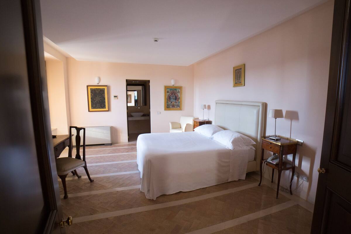 Habitación de hotel con una cama grande, una mesita de noche con lámpara, una silla, un escritorio y un radiador.