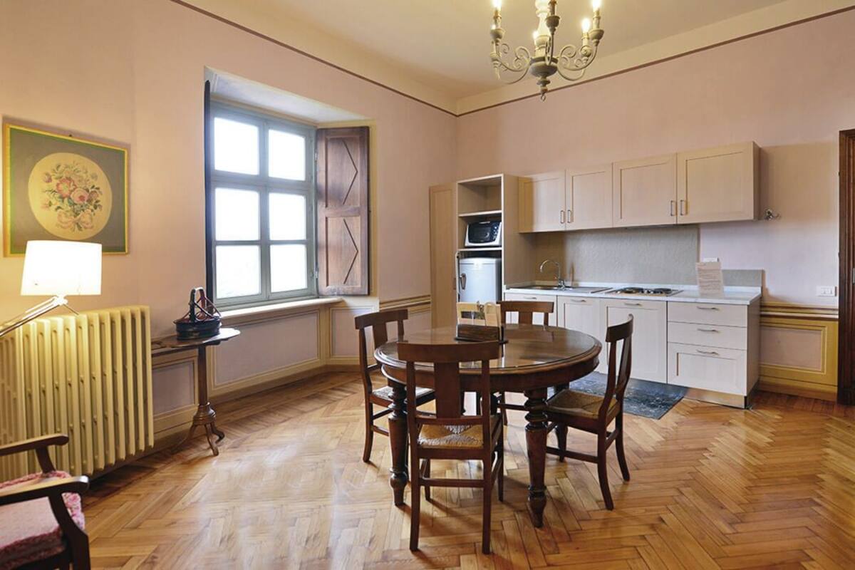 Une cuisine avec des meubles de rangement en bois, une table à manger avec des chaises, une fenêtre avec des stores et un radiateur.