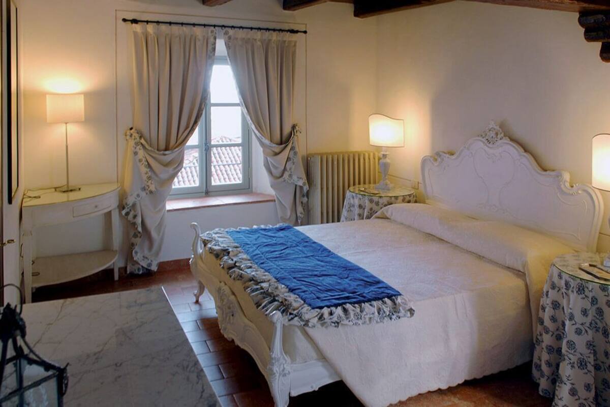 Une chambre avec un grand lit, deux tables de chevet avec des lampes, une fenêtre avec des rideaux, un radiateur et un plancher en bois.