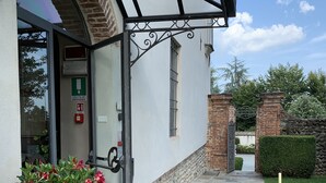 A building entrance with a glass door, a green emergency exit sign, a potted plant, and a stone pathway leading to the entrance. - Castello La Rocchetta (Sandigliano)