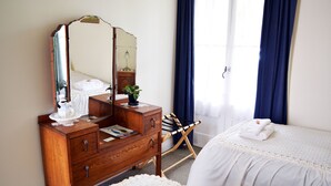 Une chambre à coucher avec une commode en bois, un miroir, un lit avec une literie blanche et une fenêtre avec des rideaux bleus.