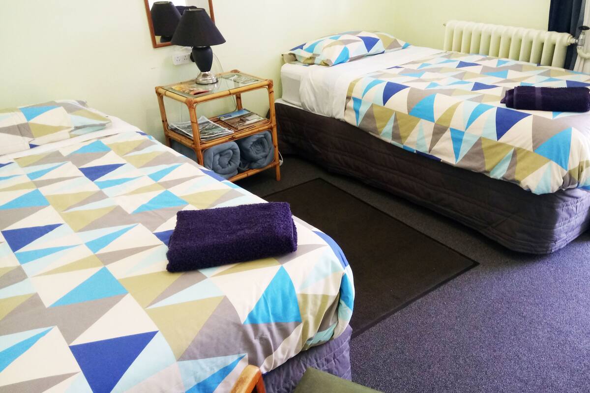 Une chambre d’hôtel avec deux lits, chacun recouvert d’une couette à motifs géométriques. On y trouve une petite table avec une lampe et un panier rempli de serviettes pliées.