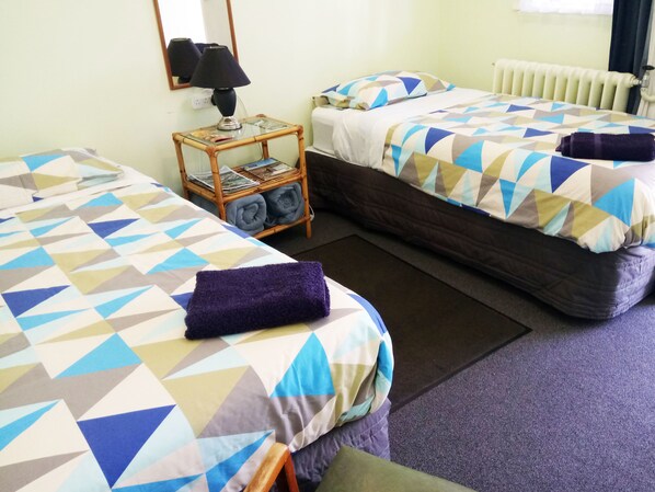 Habitación de hotel con dos camas, cada una con una colcha de diseño geométrico. Hay una mesita con una lámpara y una cesta de toallas dobladas.