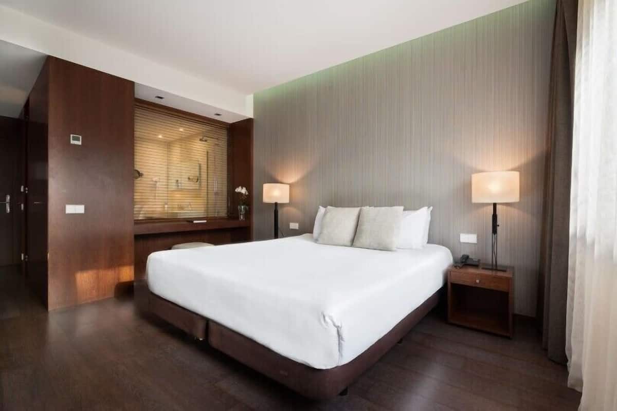 A modern hotel room with a large bed, two bedside lamps, a wooden wardrobe, and a large window with blinds.