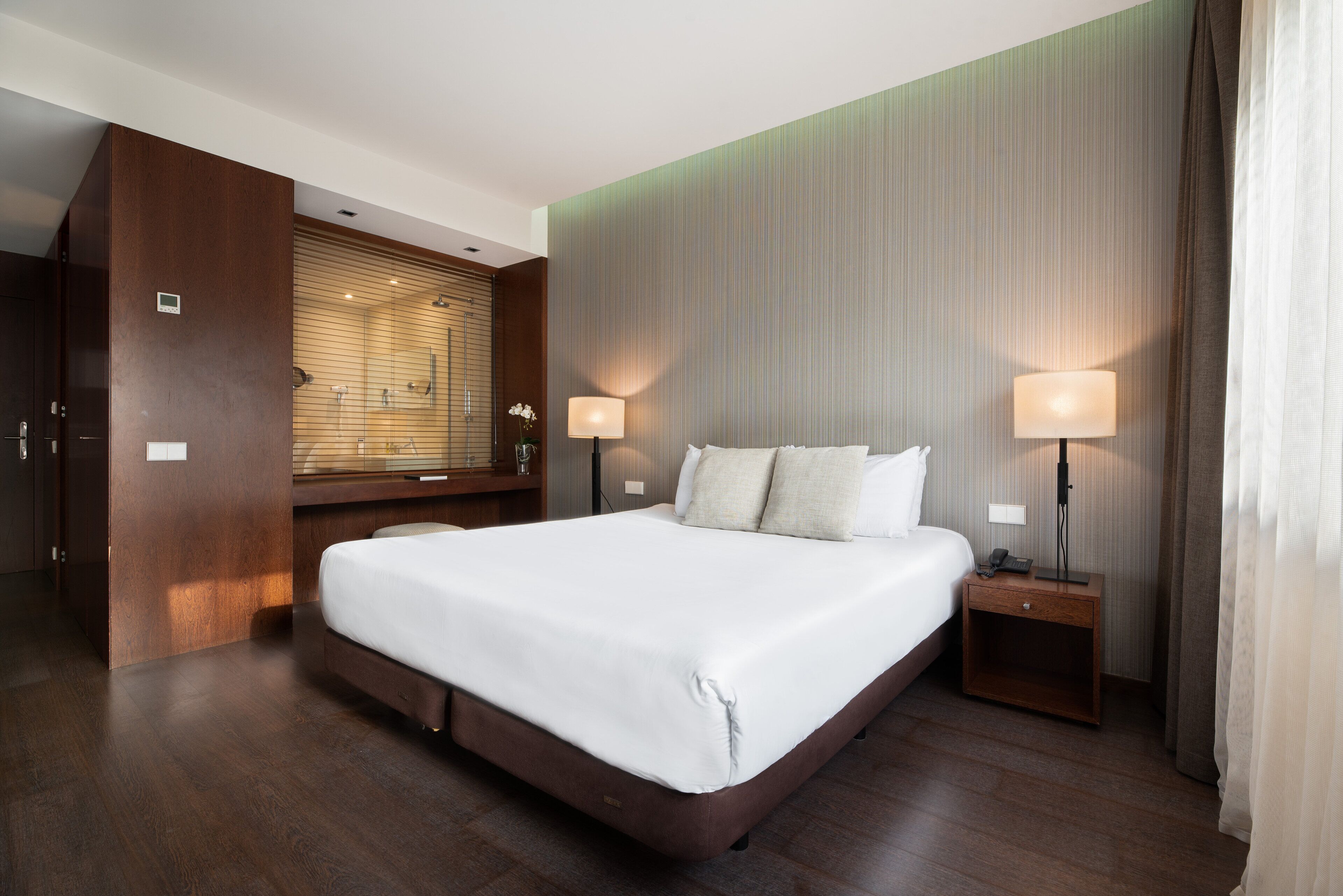 Una habitación de hotel moderna con una cama grande, cabecera de madera, dos lámparas de noche y un tocador empotrado con espejo.