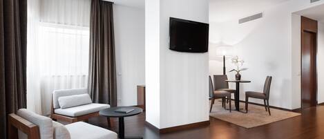 A modern living room with a sofa, a small table, a TV mounted on the wall, and a dining area with chairs and a table.