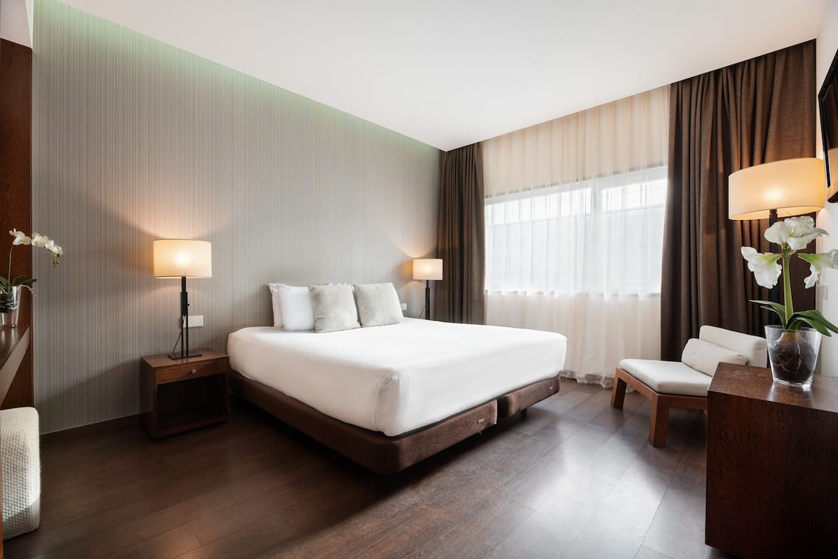 A hotel room with a large bed, two bedside tables with lamps, a chair, a desk with a lamp, and a window with curtains.