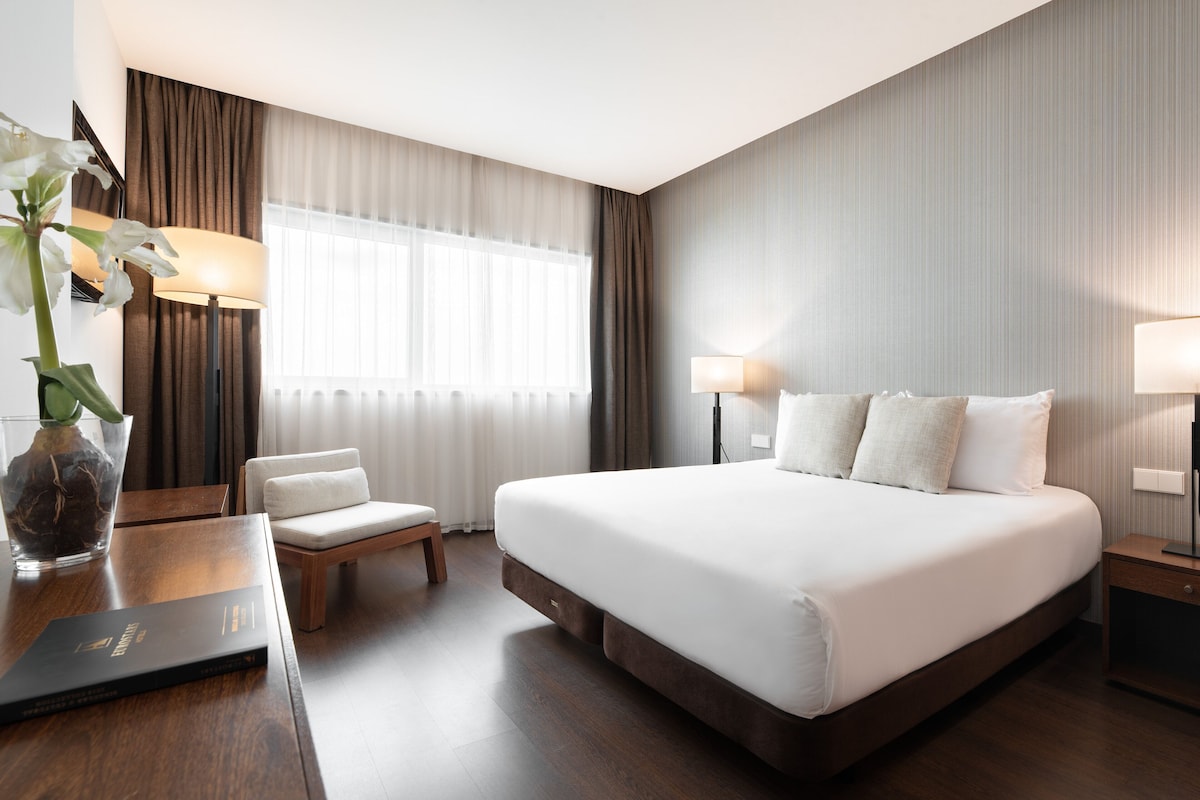 Een hotelkamer met een groot bed, een bureau met lamp, een stoel en een vaas met bloemen.