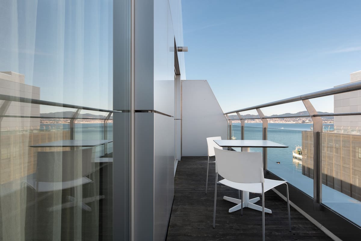 A modern balcony with a glass railing, white chairs, and a table overlooking a body of water.