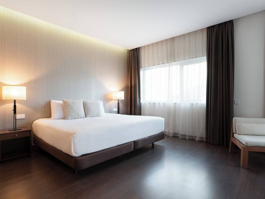 Moderni hotellihuone, jossa on suuri sänky, kaksi yöpöytää valaisimineen, tuoli ja suuri ikkuna, jossa on verhot.