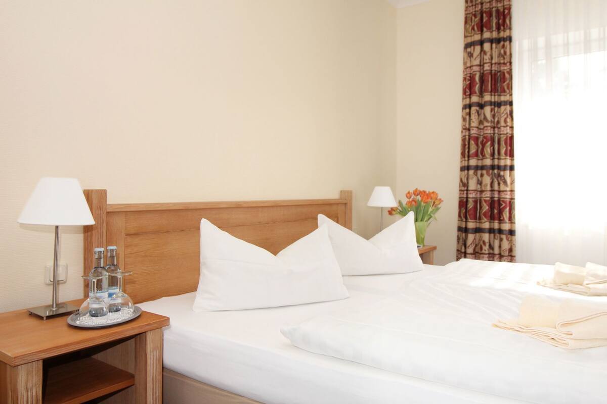 Ein Hotelzimmer mit einem hölzernen Kopfteil, zwei weißen Kissen, einem Bett mit weißer Bettwäsche, einem Nachttisch mit Lampe und einem Tablett mit Gläsern sowie einem Fenster mit Vorhängen.