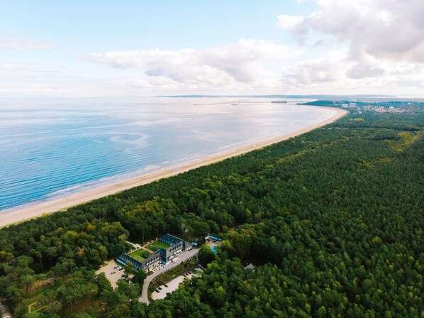 Luftaufnahme einer Küstenregion mit Strand, dichtem Wald und einem Gebäudekomplex mit mehreren Gebäuden und Parkplätzen.