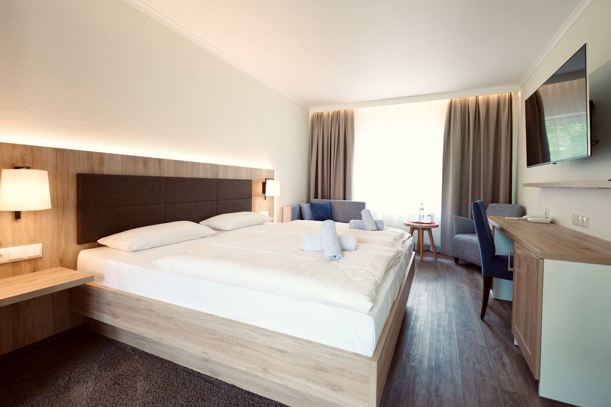 Ein Hotelzimmer mit einem großen Bett, einem Schreibtisch, einem Stuhl, einem Fernseher und einem Fenster mit Vorhängen.