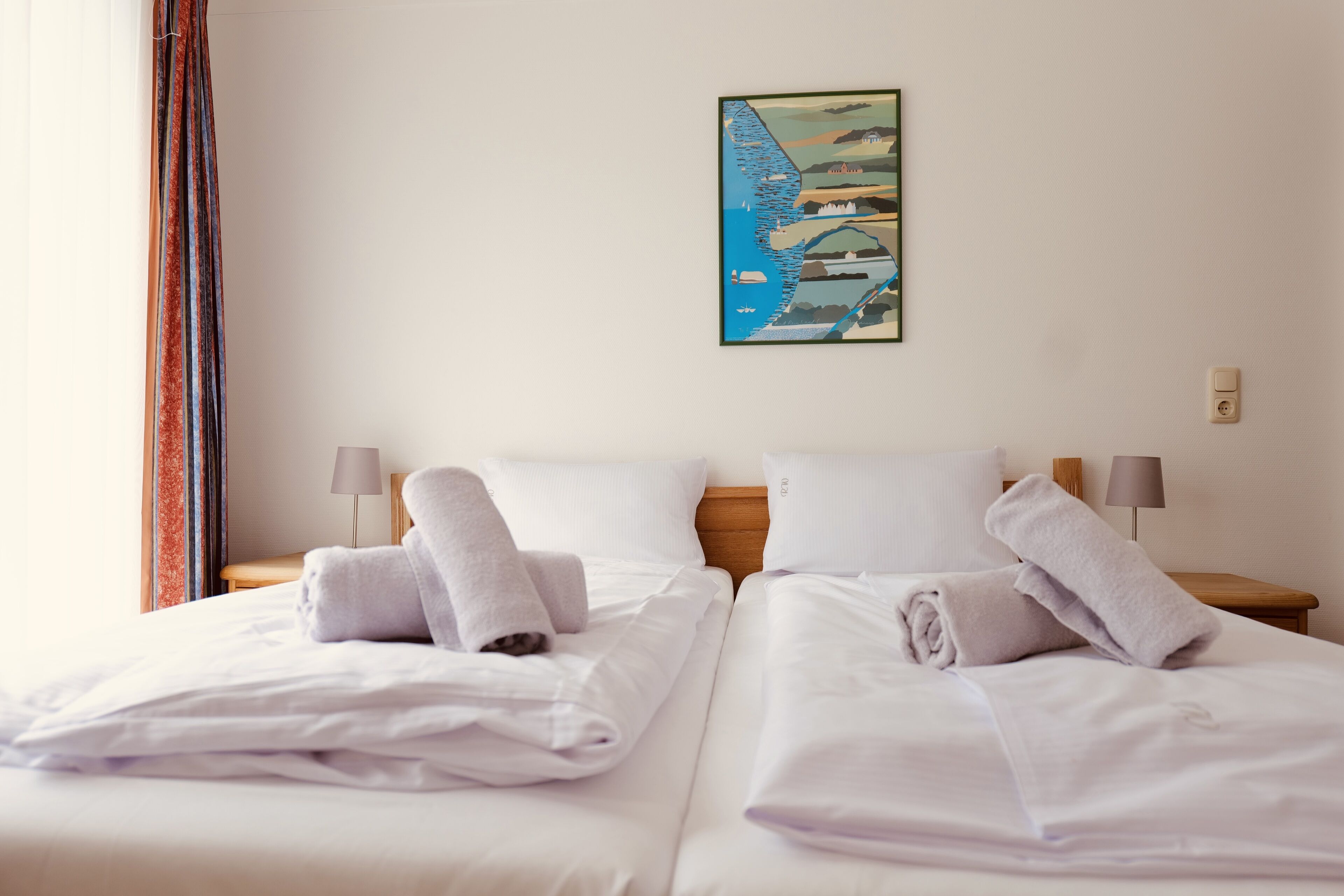 A hotel room with two beds, white linens, and towels. There is a painting on the wall depicting a coastal scene.