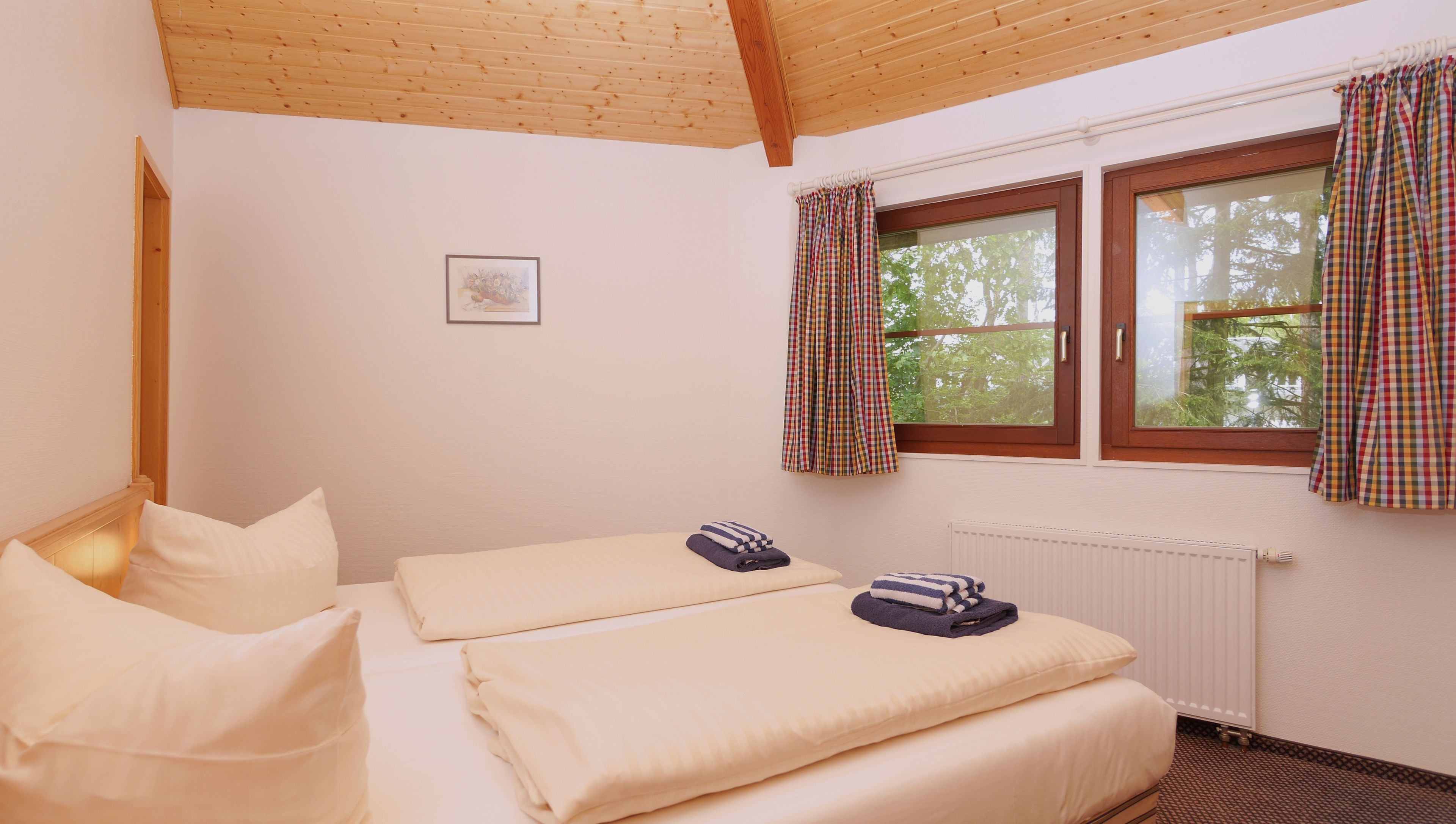 A bedroom with two beds, a window with plaid curtains, a radiator, and a framed picture on the wall.