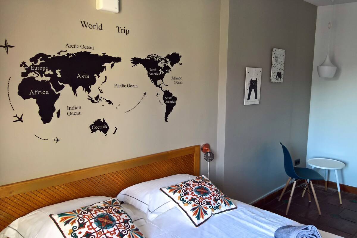 Habitación de hotel con un mural de un mapa del mundo, una cama con almohadas estampadas, una mesita y una silla.