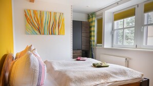 A bedroom with a bed, pillows, a colorful abstract painting, a radiator, and a window with blinds. - Ferienwohnung Fricke (Dresden)