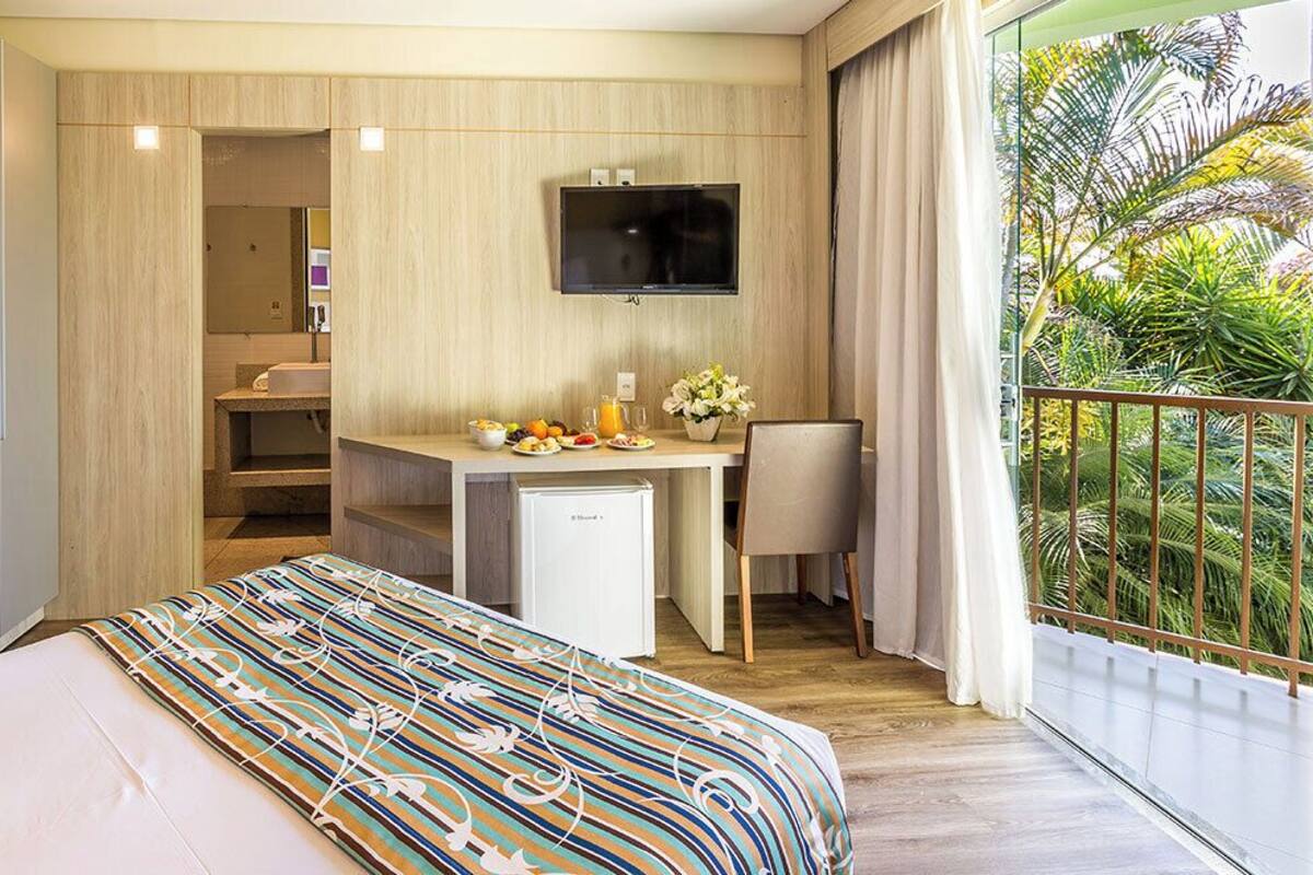 A hotel room with a bed, a desk with a computer, a TV, and a balcony with a view of palm trees.