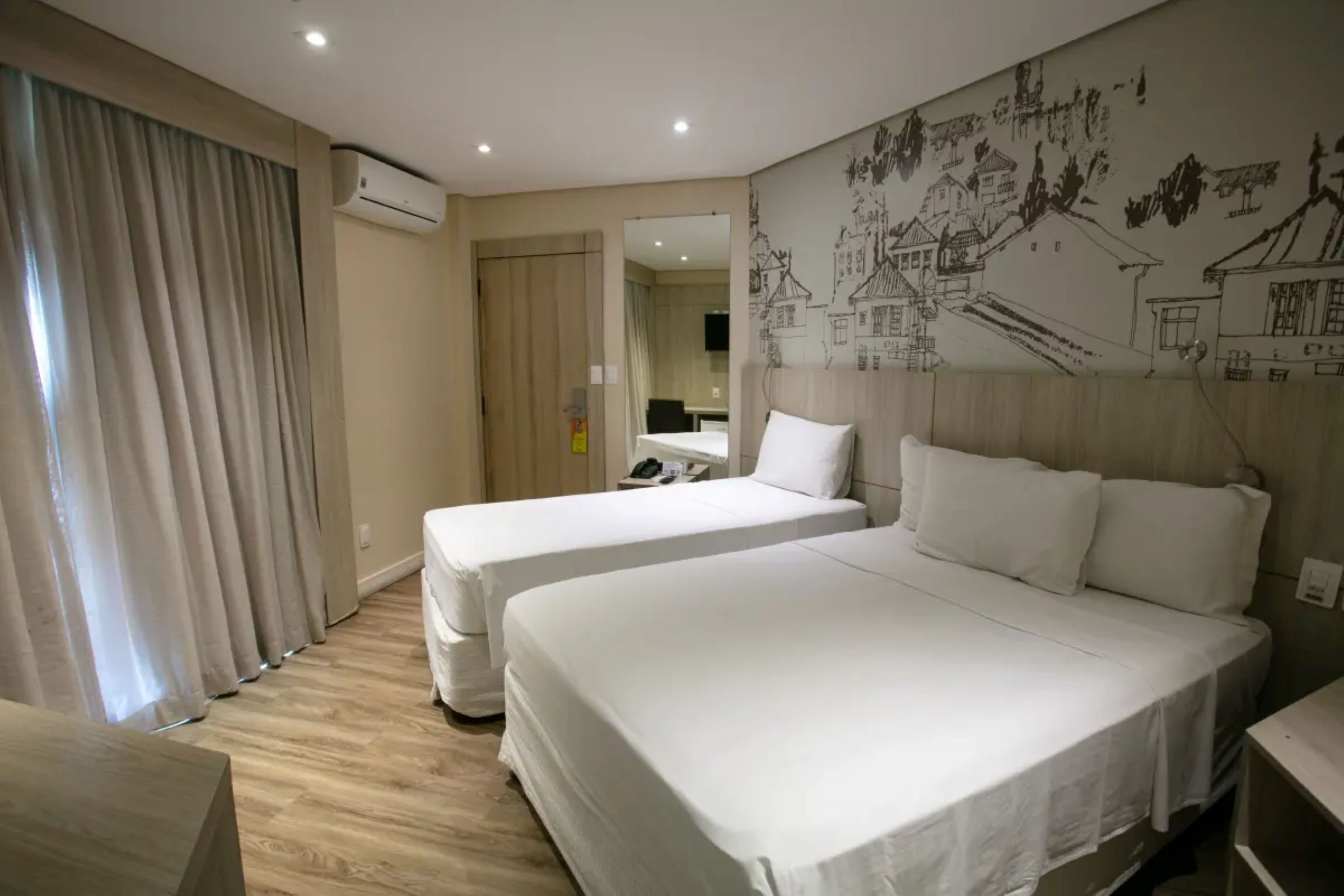 A hotel room with two beds, a wooden floor, an air conditioning unit, a wall with a mural, and a bathroom visible through a door.