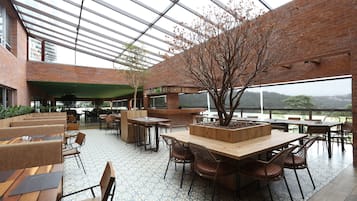 A spacious indoor area with a large wooden table surrounded by chairs, a central tree planter, and a view of the outdoors through a glass ceiling.