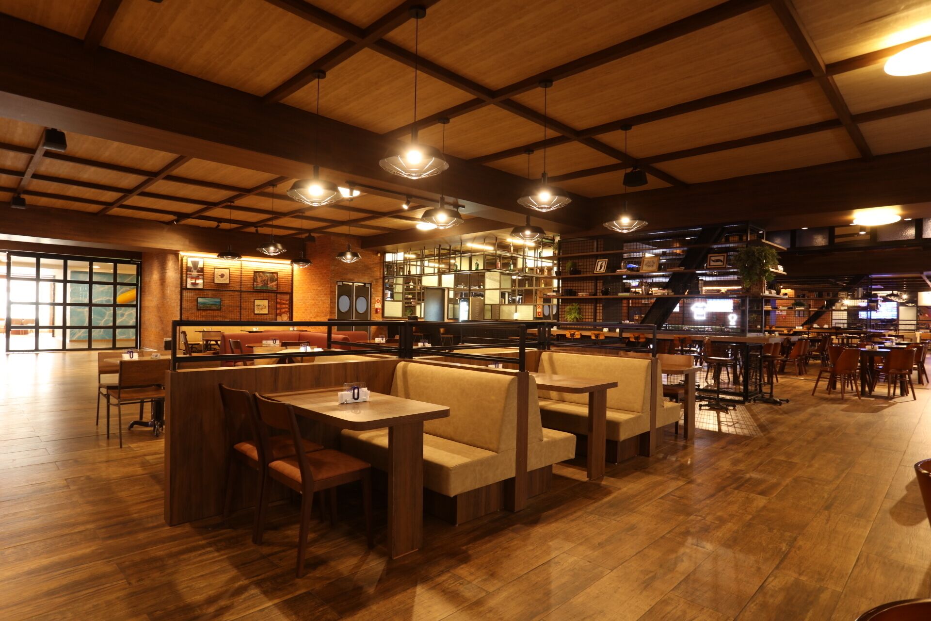 Un restaurante con suelo de madera, mesas con cabinas e individuales. Se observan luces colgantes y una barra al fondo.