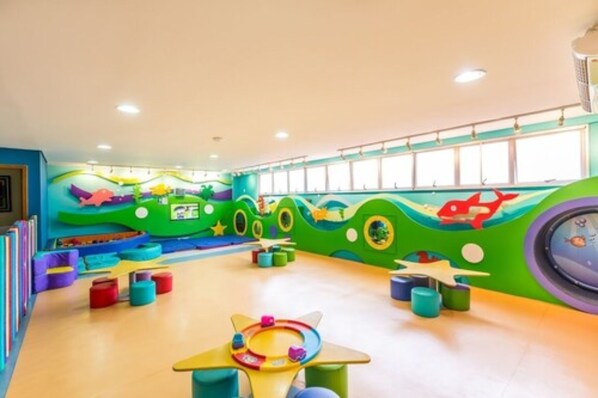 A brightly colored playroom with a wave-themed wall, colorful play structures, and a large window. - Tauá Resort Caeté (Caete)