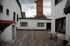 A courtyard with a tiled floor, white walls, and a brick chimney. There are two tables and chairs outside a building. - Hotel - Zum goldenen Stern (Unterspreewald)